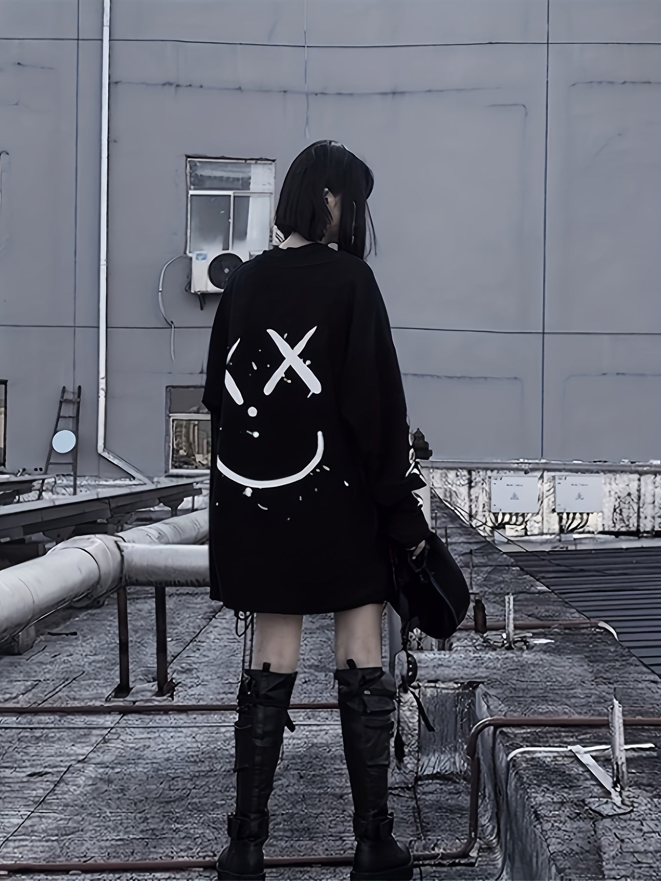 A person with black hair is standing on a rooftop, wearing black knee-high boots and a large Maramalive™ Graffiti Print Crew Neck T-Shirt, Casual Loose Long Sleeve Top For Spring & Fall, Women's Clothing with a smiley face design on the back. The background is a grey building wall.