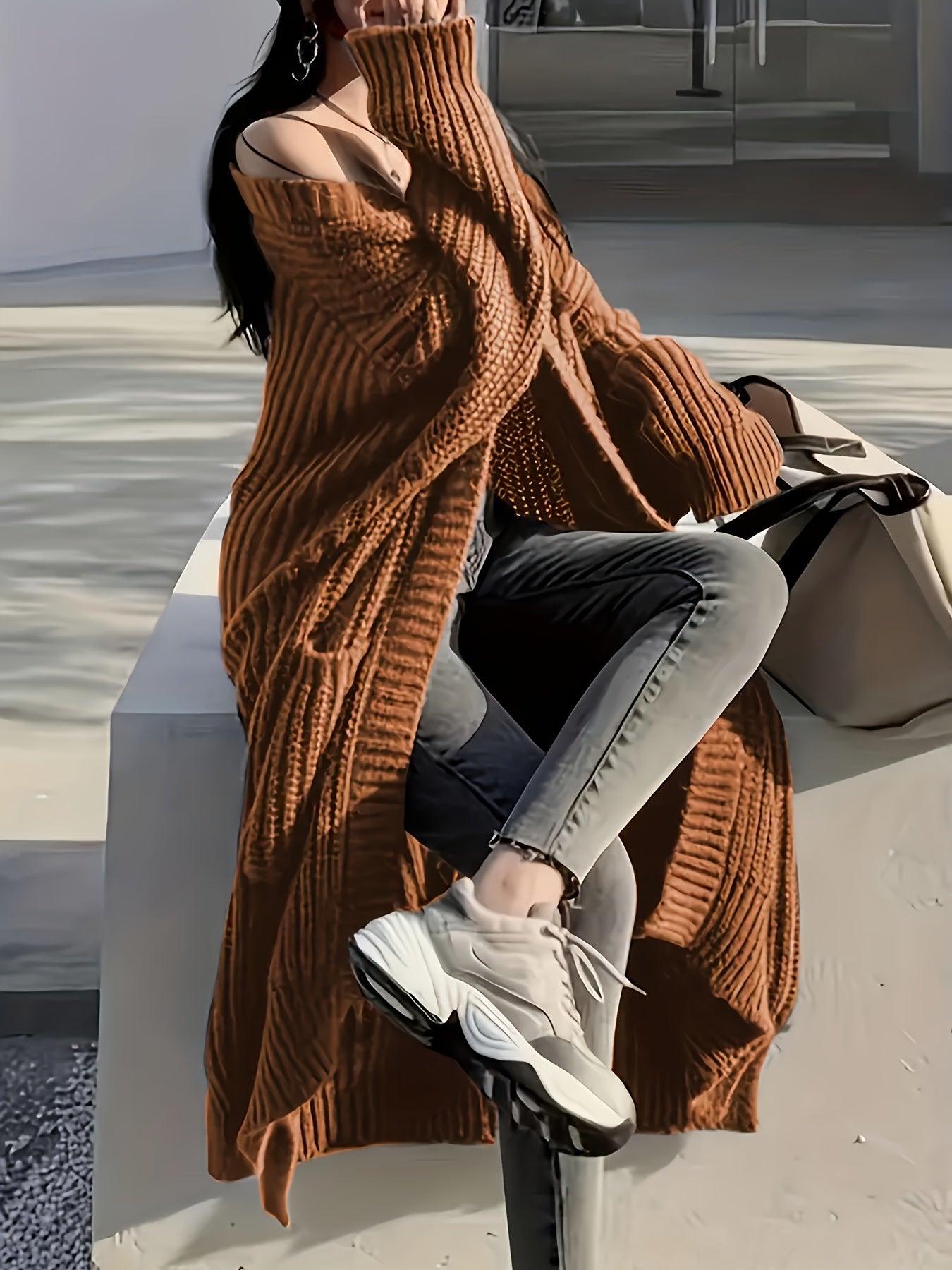 A person wearing a Maramalive™ Plus Size Open Front Loose Knit Cardigan, Casual Long Sleeve Long Length Cardigan With Pocket, Women's Plus Size Clothing, gray jeans, and white sneakers is sitting on a low wall outdoors, with a large beige bag beside them. Their face is partially covered by the cardigan.