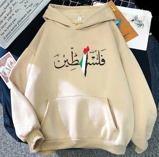 A beige winter hooded pullover is displayed on a bed. The Maramalive™ Autumn And Winter Fleece Warm Hoodie Jacket Casual Sweatshirt, a polyester men's hoodie, features Arabic calligraphy and a design incorporating the colors of the Palestinian flag on the front. Various papers and items are scattered around it.