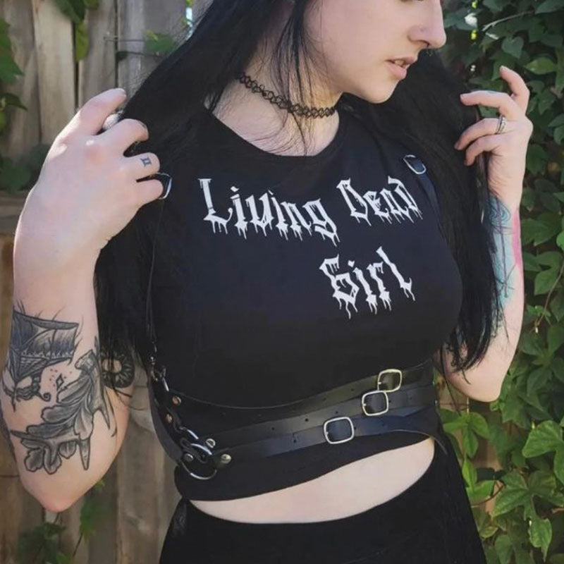 A person wearing a black Maramalive™ Gothic Style Printed Top Short Sleeve with "Living Dead Girl" written on it, a black choker necklace, and a black harness belt. They have tattoos on their arms and are standing outdoors with greenery in the background.