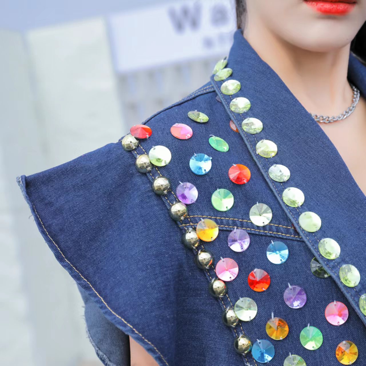 Close-up of a person wearing a Maramalive™ Heavy Duty Diamond Studded Denim Vest With Wooden Ear Edge with colorful, round reflective studs and gold beads. The blue coat features beaded sequins that catch the light. The person is also wearing a silver necklace and has bright red lipstick.