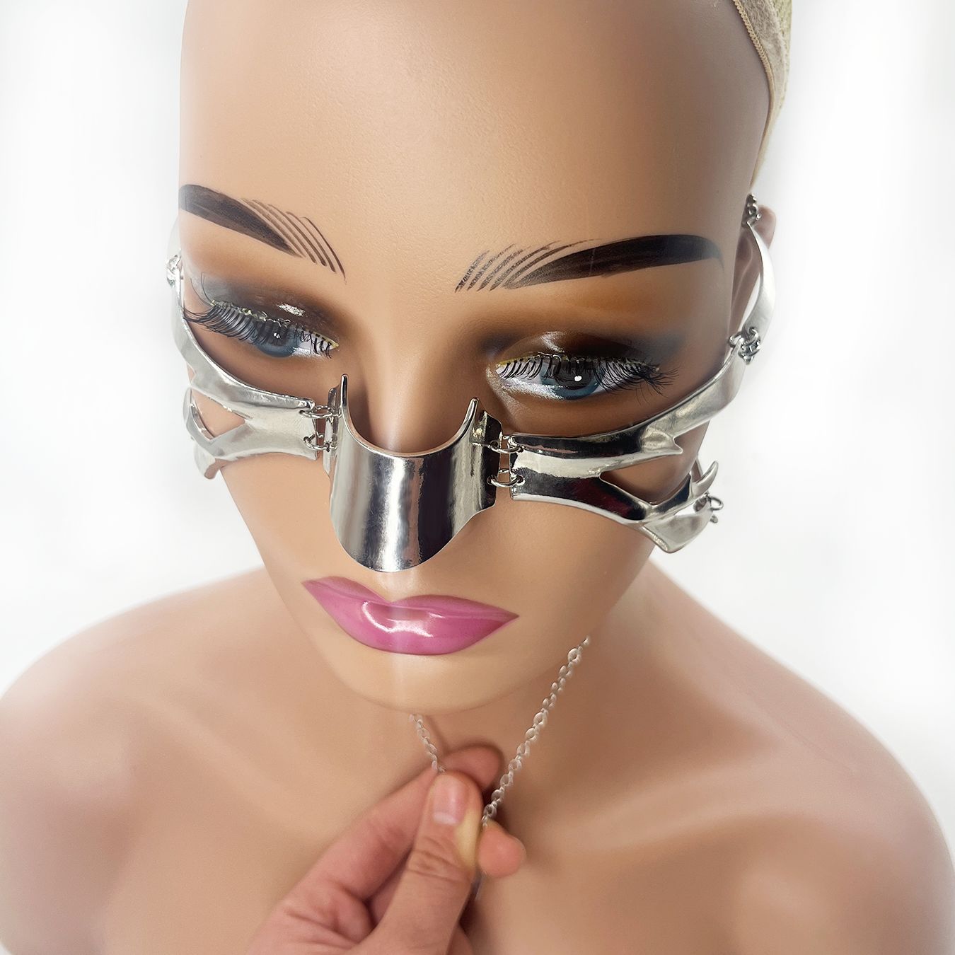 A mannequin head wears a metallic facial accessory, like an intricate alloy mask, covering the nose and cheeks. A hand is holding a chain attached to it. The Adjustable Irregular Fluid Lip Ring Mask from Maramalive™ features multiple segments and hinges, perfect for dressing up in a unique style.
