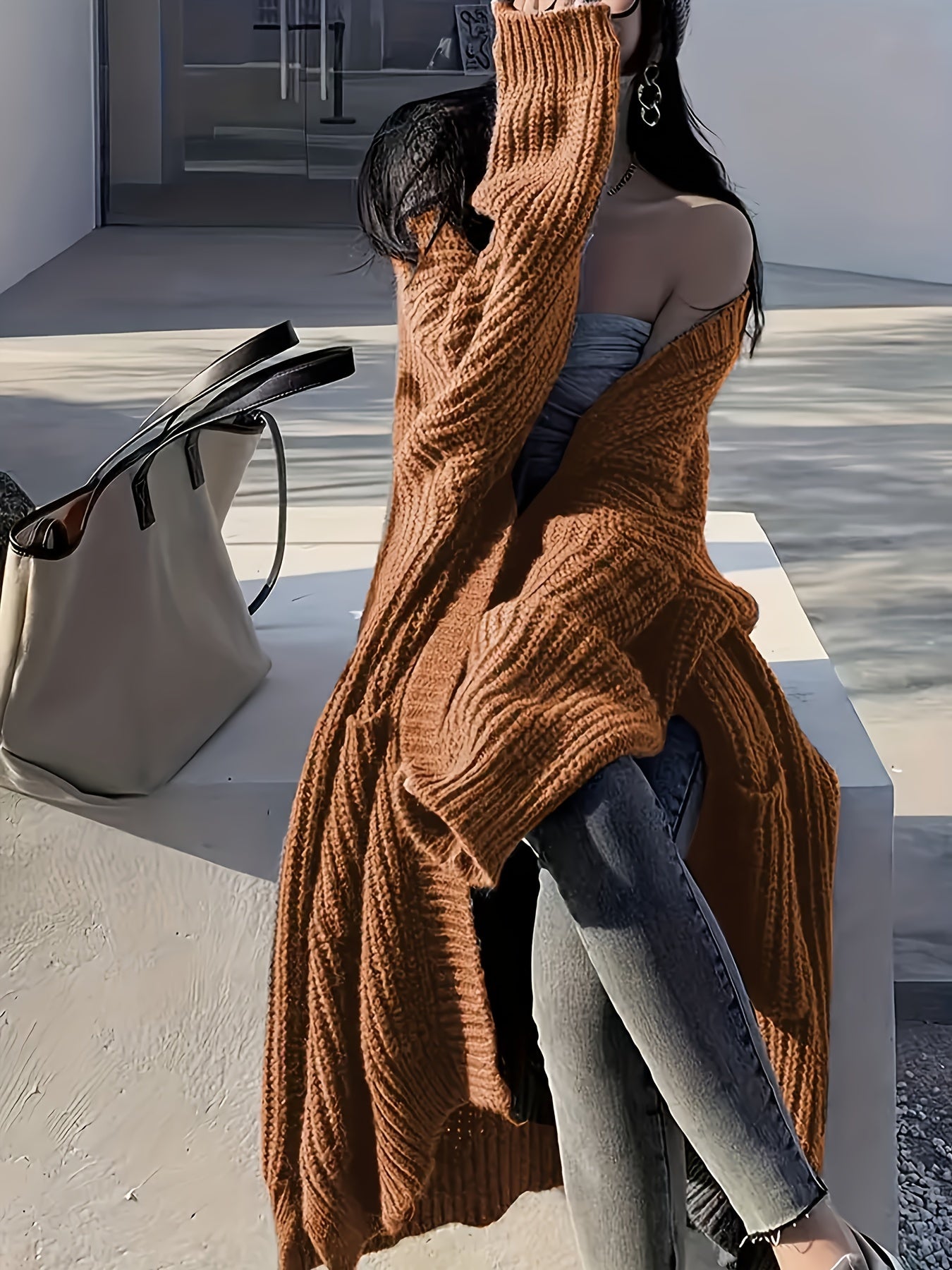 A person in a Plus Size Open Front Loose Knit Cardigan, Casual Long Sleeve Long Length Cardigan With Pocket, Women's Plus Size Clothing by Maramalive™, sits on a concrete surface next to a beige handbag.