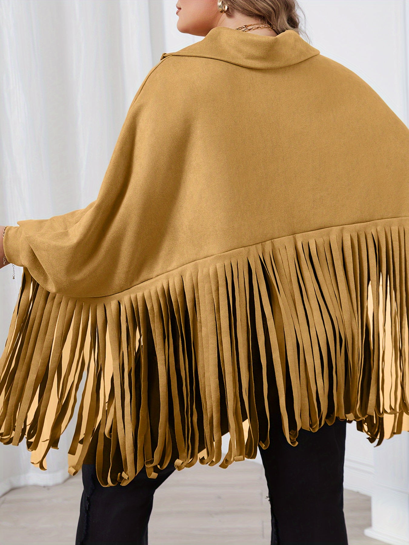 A person wearing a mustard-colored, fringed Maramalive™ Women's Plus Solid Batwing Sleeve Mock Neck Fringe Trim Cloak Top with batwing sleeves, viewed from the back, with white curtains in the background. Perfect for Fall/Winter.