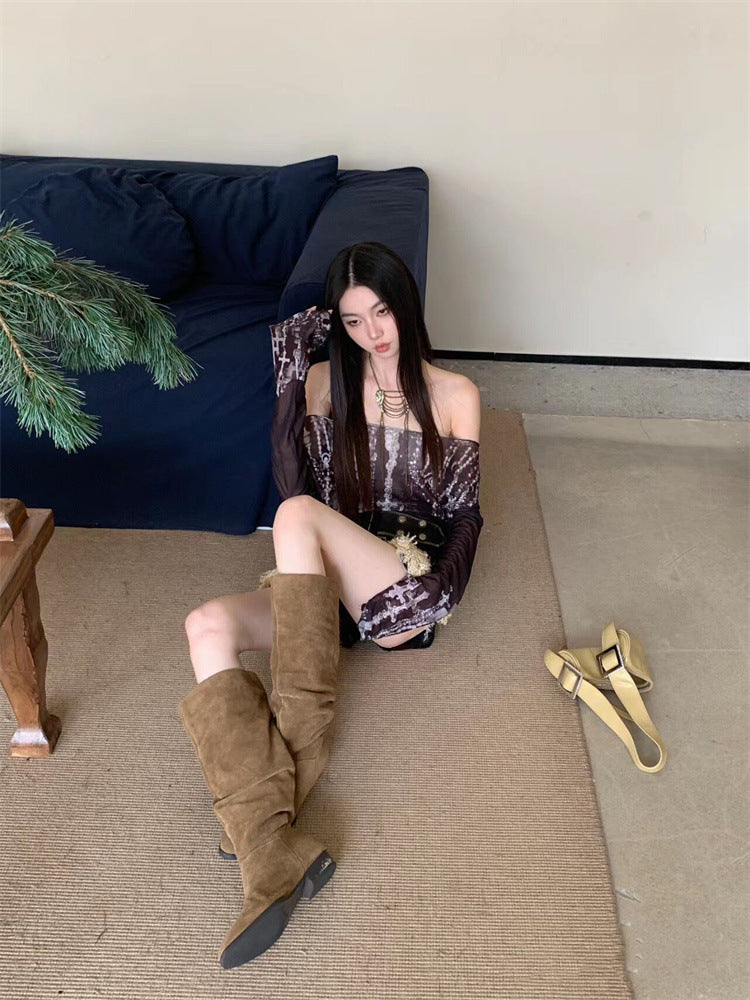 A woman with long, dark hair sits on the floor leaning against a navy sofa, wearing a **Maramalive™ Off-shoulder Printed Mesh Long Sleeve Bottoming Shirt Sun Protection Shirt** and brown knee-high boots. A slim fit beige belt lies on the floor beside her, adding to her chic urban style.